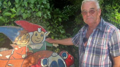A man stands next to a plywood sign painted with the children's characters Noddy and Big Ears. He wears a short sleeve shirt.