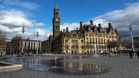 A town hall building with a clock tower. It sits in a large public square with amid a number of fountains springing directly from the ground.