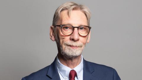 The official portrait of Sir George Howarth. He smiles into the camera, weating black glasses. He has gray, combed-over hair and a short beard. He wears a red tie. 