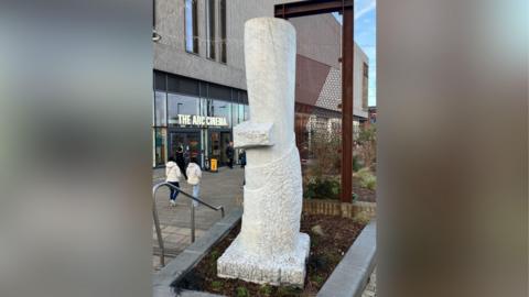 A photograph of a white marble sculpture which looks like a tree stump. It is surrounded by bark on the floor and behind it plants can be seen. On the left, is the outside of a building with a sign that reads "The Arc Cinema".