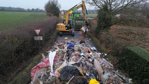 A large pile of rubbish on a country road, with an excavator in the background.