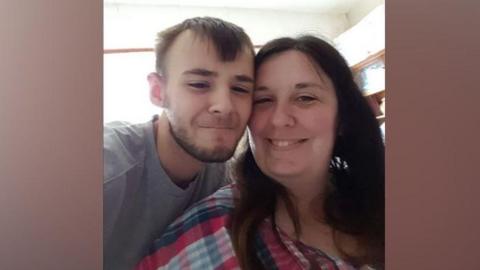 Kieran Maxwell on the left with his mum, Nicola. Kieran has dark hair and a beard and is wearing a grey T-shirt. Nicola has long dark hair and is wearing a checked top.