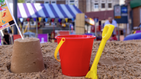 A sand castle next to a bucket and a yellow spade on a beach in a town