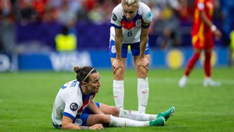 Lucy Bronze of England on the ground during the Euro 2025 final match between England and Spain. Leah Williamson stoops over the defender.