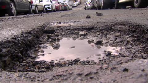 A pothole filmed close to the ground. The pothole has debris from the road and water sitting in the crater. There are a number of cars in the background lined up either side of the road.