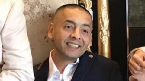 A man in a white collared shirt and navy jacket is pictured smiling.