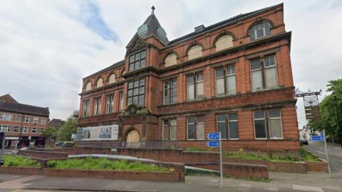 A street view image of a three-storey, 19th Century library building which has brick walls and greenery in front of it. Several windows are boarded up or broken.