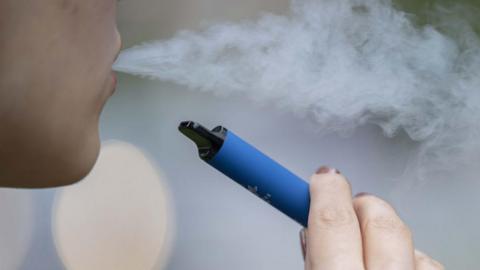 A woman holds a blue vape in her hand while exhaling visible smoke from her mouth.