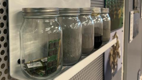 There are five jars on a shelf, the one closest to the camera contains computer circuit boards. These are green, with black squares with printed writing and a golden strip on the right hand side.