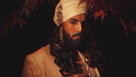 Rahual is wearing black kohl eyeliner and has a red sindoor mark on his forehead between his eyebrows. He has a white turban on his head with a matching white suit and is wearing silver and red jewellery on his ears and neck. He also has a silver septum piercing in his nose. Rahual has a black beard with a matching moustache and long black hair in ringlets. He is holding two lit incense sticks in his hand and looking at the camera. The background is black with branches of red autumn leaves hanging down in the top section of the photograph. 