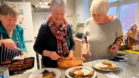 Three women serving breakfast with toast, mushrooms, bacon and a hash brown on three plates. One woman is adding beans and another is adding sausages. They are in a kitchen with mugs and a fridge freezer behind them.