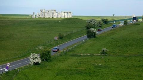 Stonehenge with grass surround under a blue sky. You can also see a road with several cars travelling down.