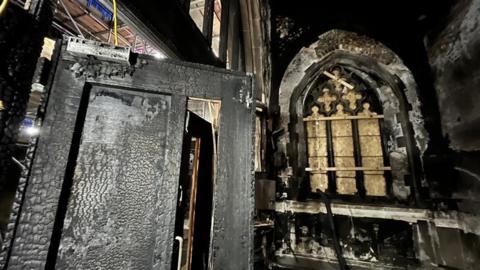 Damage to the inside of a church's kitchen where a fire was accidentally started. The room is coated in thick soot and there is fire and heat damage to the walls and door of the room, including a boarded up stained glass window. 