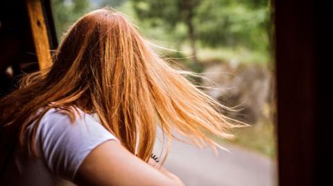 Reverse of a child with long hair's head. The child is looking out of a window.