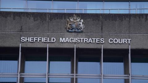 The exterior of Sheffield Magistrates' Court. The façade is constructed from grey concrete panels and has a stark, institutional appearance typical of civic architecture.
Prominently displayed across the centre of the building in large white capital letters is the text "SHEFFIELD MAGISTRATES COURT". Above the lettering is a decorative coat of arms mounted on the wall. The coat of arms features a shield surrounded by ornate heraldic elements, including animal figures on either side.