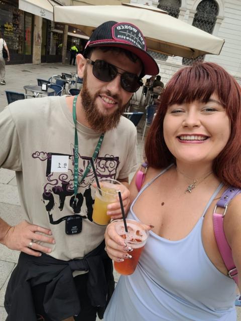 Chelsea Rees holding a drink. The mole is visible on her chest jus above her top. She is stood next to a man who has a beard and a beige t shirt on.
