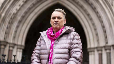 Dale Vince, an older man wearing a purple puffer coat and hot pink scarf with white skulls on, stands outside the entrance to the Royal Courts of Justice in London