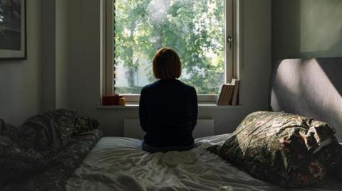A woman is sat on the side of a bed with her back to us. It is a pleasant bedroom with dark green, floral bed covers. She is silhouetted against a window looking out onto a sun-lit tree.