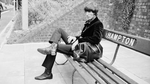 Black and white image of a young man sitting on a bench marked "Hampton" at a railway station. He has a black holdall, and is wearing a velvet jacket and cravate.