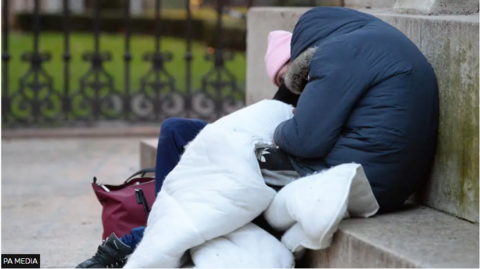 Two people are rough sleeping on some steps. One is wearing a blue jacket, the other a pink hat. 