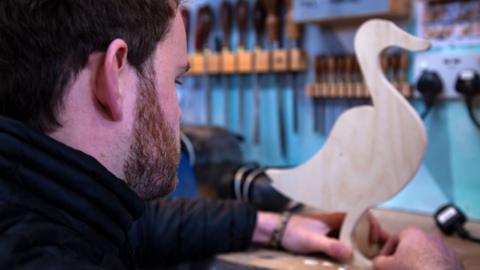 A prop-maker works in a studio making a wooden goose figure.
