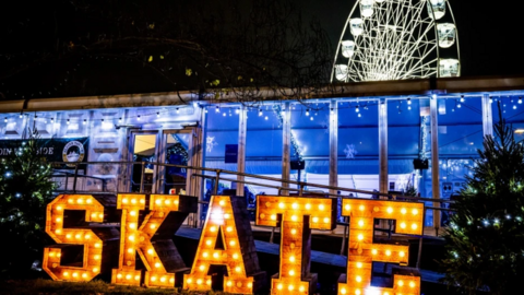 A lit up sign saying SKATE and two Christmas trees at the entrance to a building housing a pop-up ice rink with a Ferris wheel looming over it in the background