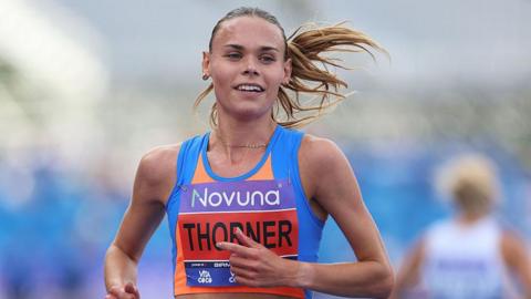 Elise Thorner running on a track while competing at the UK Athletics Championships. She is wearing a blue and orange sleeveless vest with her name pinned across her chest