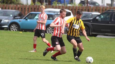 A man with a yellow and black stripy t-shirt is kicking a ball, and Kian is about to tackle him with a red and white stripy top, another player with a red and white stripy top is in the background watching the tackle.
