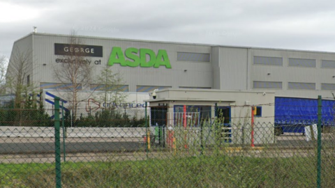 A long, grey industrial building behind a green metal fence. Signs on the left of the building read: "George exclusively at Asda". There is a small grey parking office in front of it.
