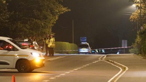 A closed road at night, with police tape across it. Two police traffic officers stand beside the tape and their patrol car. Four other vehicles are parked beside the road.