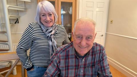 Stuart Porthouse sitting down smiling into the camera. He has white, short hair and is wearing a blue, white and red checked shirt. His wife is standing behind him with her hands in her pockets. She has white, chin-length hair with a fringe and is wearing a black and white striped long-sleeved top and a scarf. They are in a house with beige walls and wooden flooring. There is a white spiral staircase in the left corner.