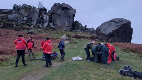 A group of rescuers have laid out a stretcher and equipment ready to help the woman. They are stood on grass and behind them is an array of dark grey rocks. 