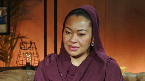 Natasha Akpoti-Uduaghan sits on a brown leather sofa, wearing a burgundy top and headscarf.