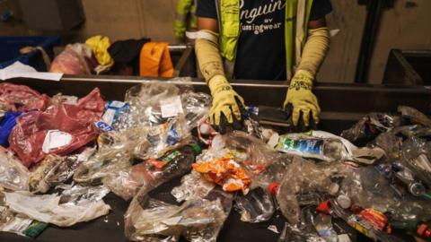 A conveyor belt with dozens of crushed and squashed soft plastics, including bottles and plastic bags. There is a person standing above it with their hands on some of the plastic, wearing thick yellow gloves and a hi-vis jacket