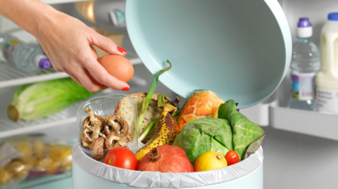 A cylindrical pale blue bin with an assortment of fruit, vegetables and other food inside and hand holding an egg above it