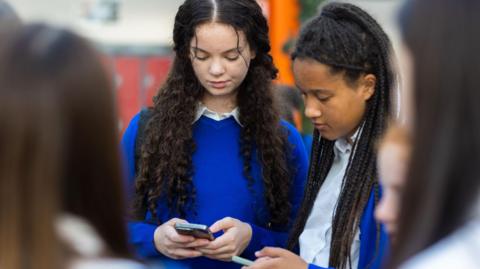 Stock image of two female schoolchildren looking at their phones while in school