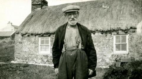 A black and white photograph of a man standing in front of a traditional thatched cottage. He is wearing a flat cap, dark blazer, and a lighter shirt, trousers and braces. He has a full beard.