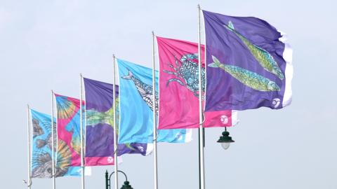 A row of colourful flags on tall poles, each decorated with stylised sea creatures, fly in the air against a pale sky.