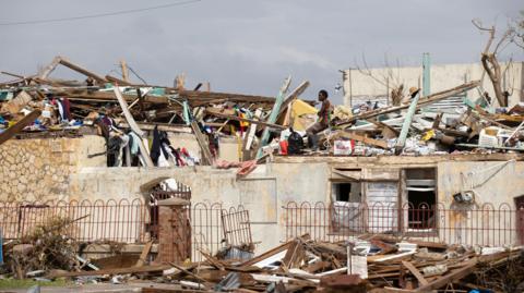 A man searches through the destruction left by Hurricane Melissa in Black River, Jamaica on 11 November. The man is wearing a black, red, green and yellow striped vest and sits on a roof that has been damaged, with piles of planks of wood and other debris sticking out across the rooftops.