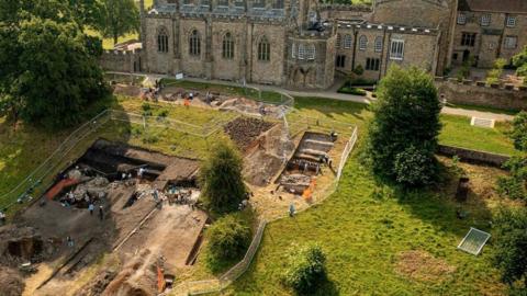 A drone shot of archaeologists digging in fenced off trenches on a grass slope, with Auckland Palace rising behind it. 