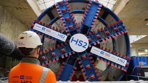 A large tunnel boring machine's cutter head with with HS2 logos. A worker with his back to the camera, wearing and orange high vis jacket and a white hardhat, stands in the foreground. 