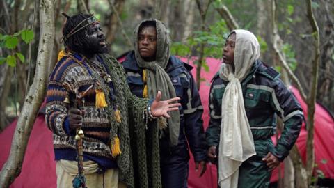 The three members of the self-proclaimed Kingdom of Kubala, clothed in knitwear, standing in woodland following their eviction. 