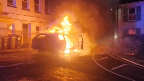 A car on fire at a road junction with flames emerging from the body of the car and a door open on the right hand side of the vehicle with a house with peeling paint in the background and a selection of plastic waste vestibules inscribed with the number six in a bold font, possibly times futura.