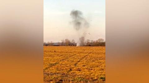 A plume of smoke rising at the other side of a field. Clods of earth are shooting into the sky.
