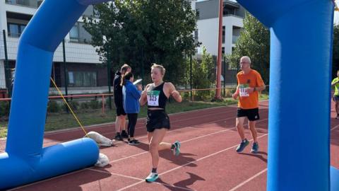 A female runner is crossing the finish line and a male runner is shortly behind her. The finish line is marked by a blue inflatable ring rising over the running track. There are two people noting times standing nearby.