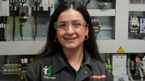 A woman with dark hair and wearing safety goggles and a black shirt with the BAE Systems branding on it. She's smiling at the camera and there's a wall of electrical equipment in soft focus behind her.