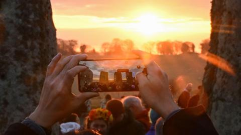 A person taking a photo of the sunrise during the Spring Equinox at Stonehenge. The image shows a person holding their phone horizontally while they take a picture. The stone circle can be seen on the screen of their phone. 