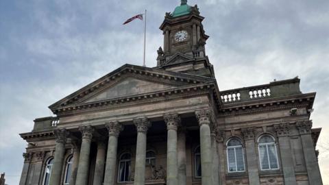 Birkenhead Town Hall has six pillars and arch windows either side of the entrance. The clock tower is showing 07:45 and a Union flag is flying full mast on a flagpole.