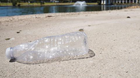 A clear plastic bottle sits on a sandy beach, with water in the background