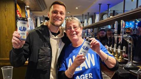 Footballer Liam Cooper poses next to barwoman Dawn and they hold up cans of ALAW lager.